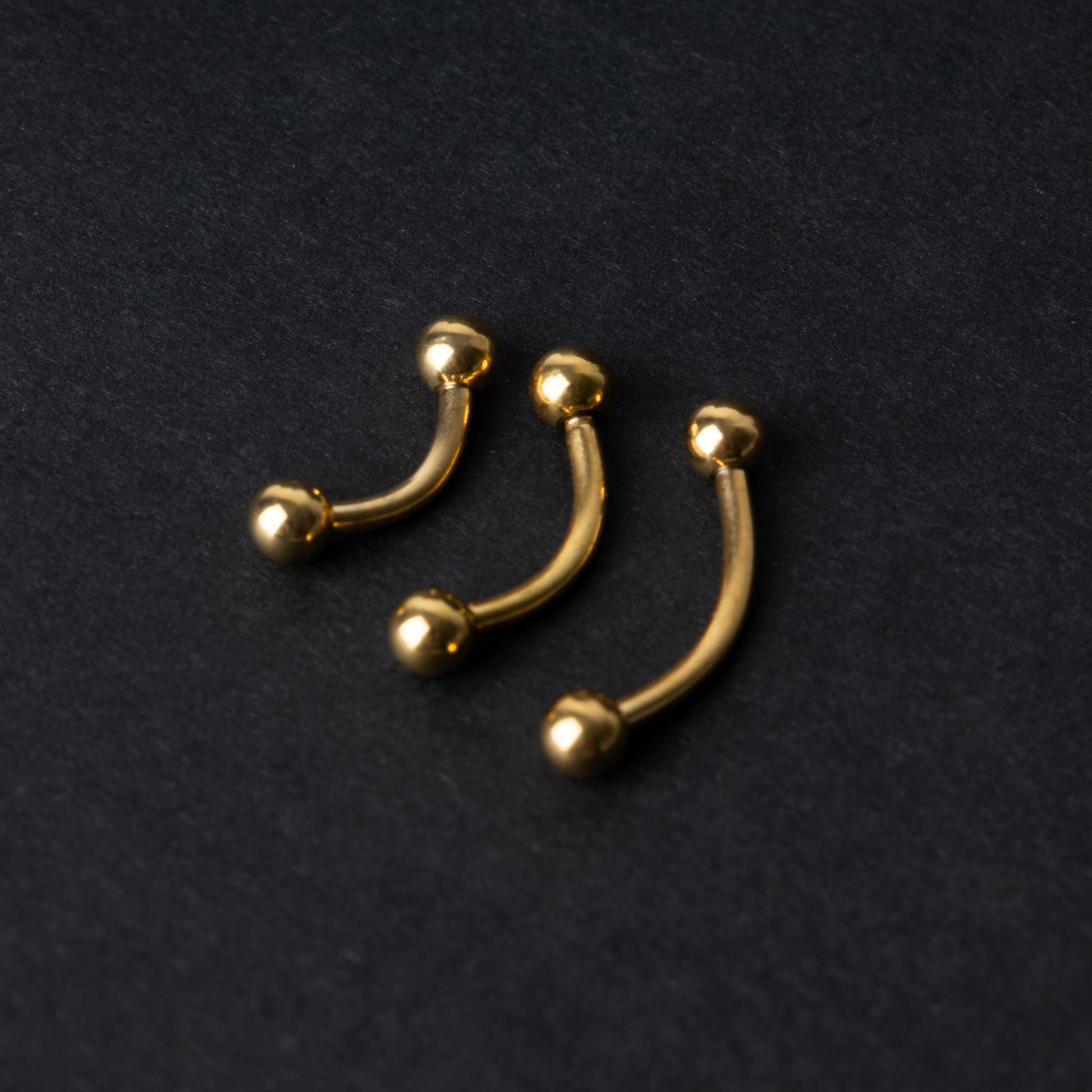 Gold curved barbells in three sizes with smooth ball ends, displayed on a dark background.