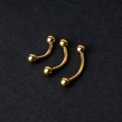 Gold curved barbells in three sizes with smooth ball ends, displayed on a dark background.