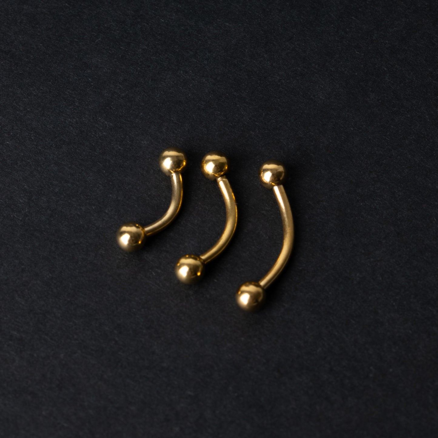 Gold curved barbells in three sizes with smooth ball ends, displayed on a dark background.