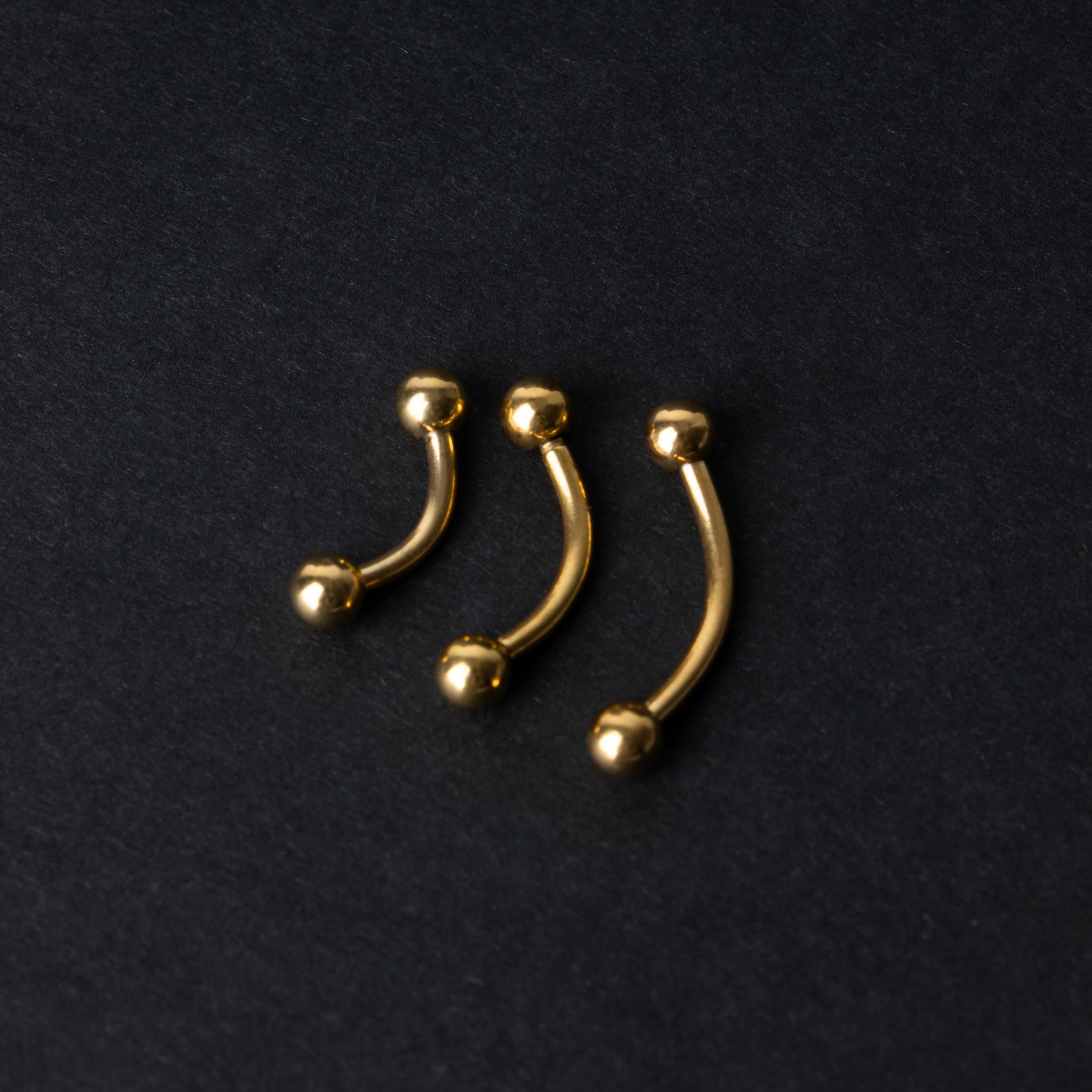 Gold curved barbells in three sizes with smooth ball ends, displayed on a dark background.