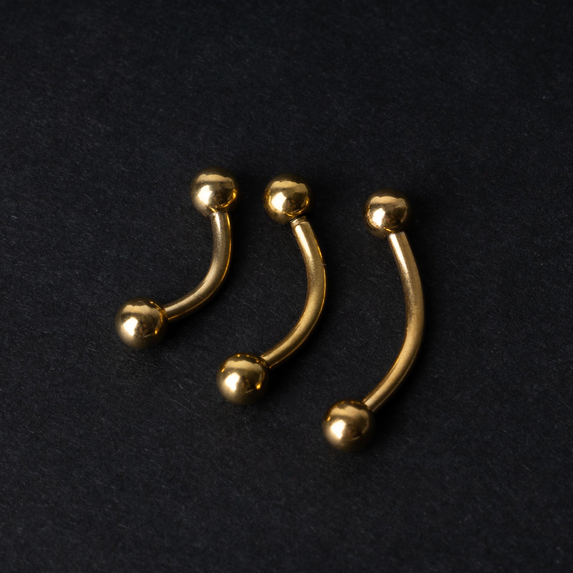 Gold curved barbells in three sizes with smooth ball ends, displayed on a dark background.