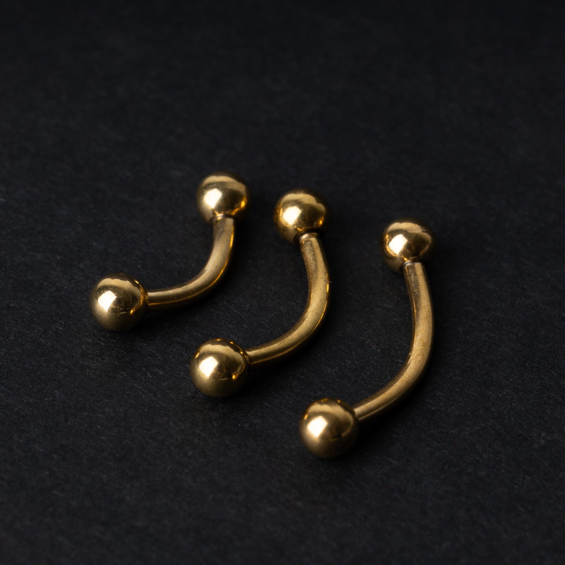 Gold curved barbells in three sizes with smooth ball ends, displayed on a dark background.