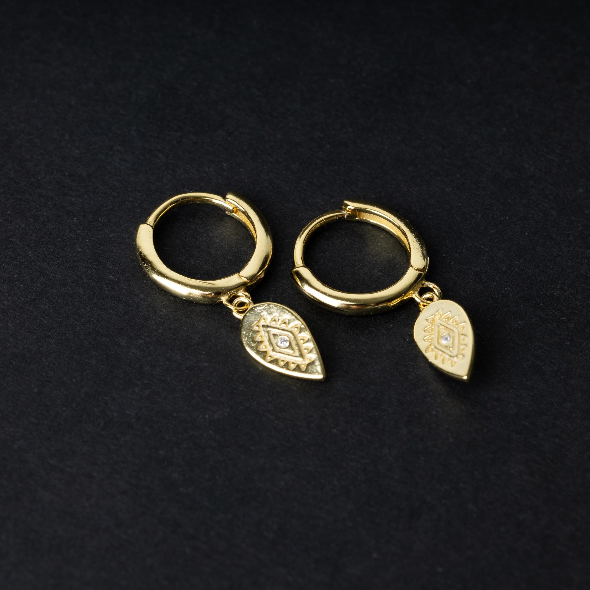 Gold hoop earrings with detailed teardrop-shaped charms, displayed on a dark background.