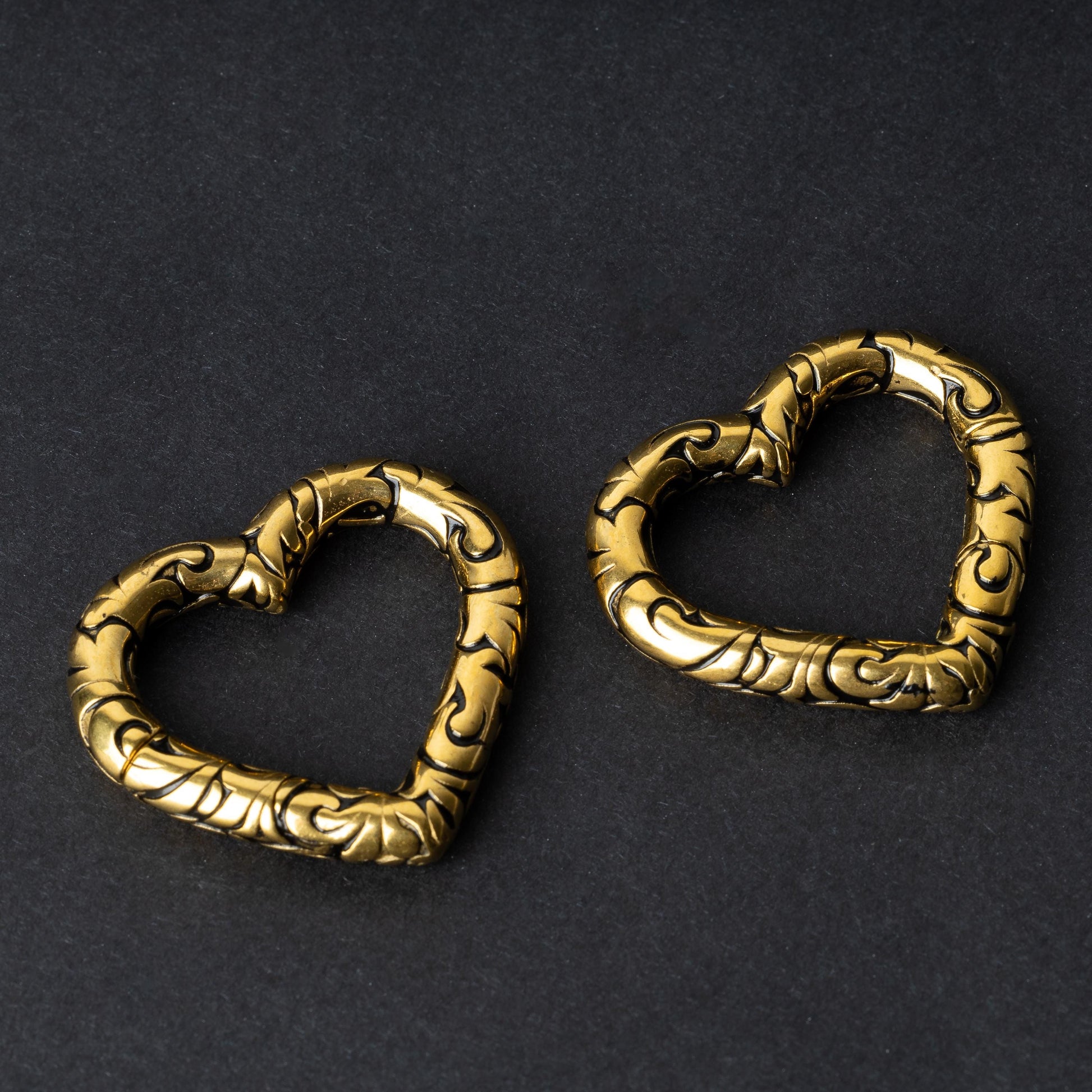 Two heart-shaped engraved saddle plugs in gold on a dark background.
