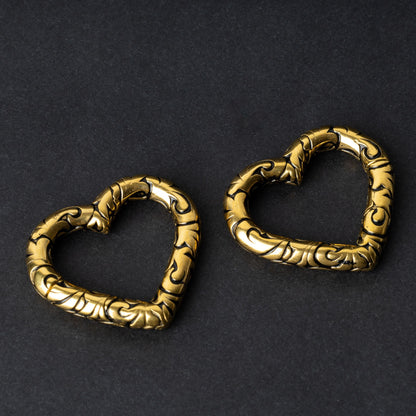 Two heart-shaped engraved saddle plugs in gold on a dark background.
