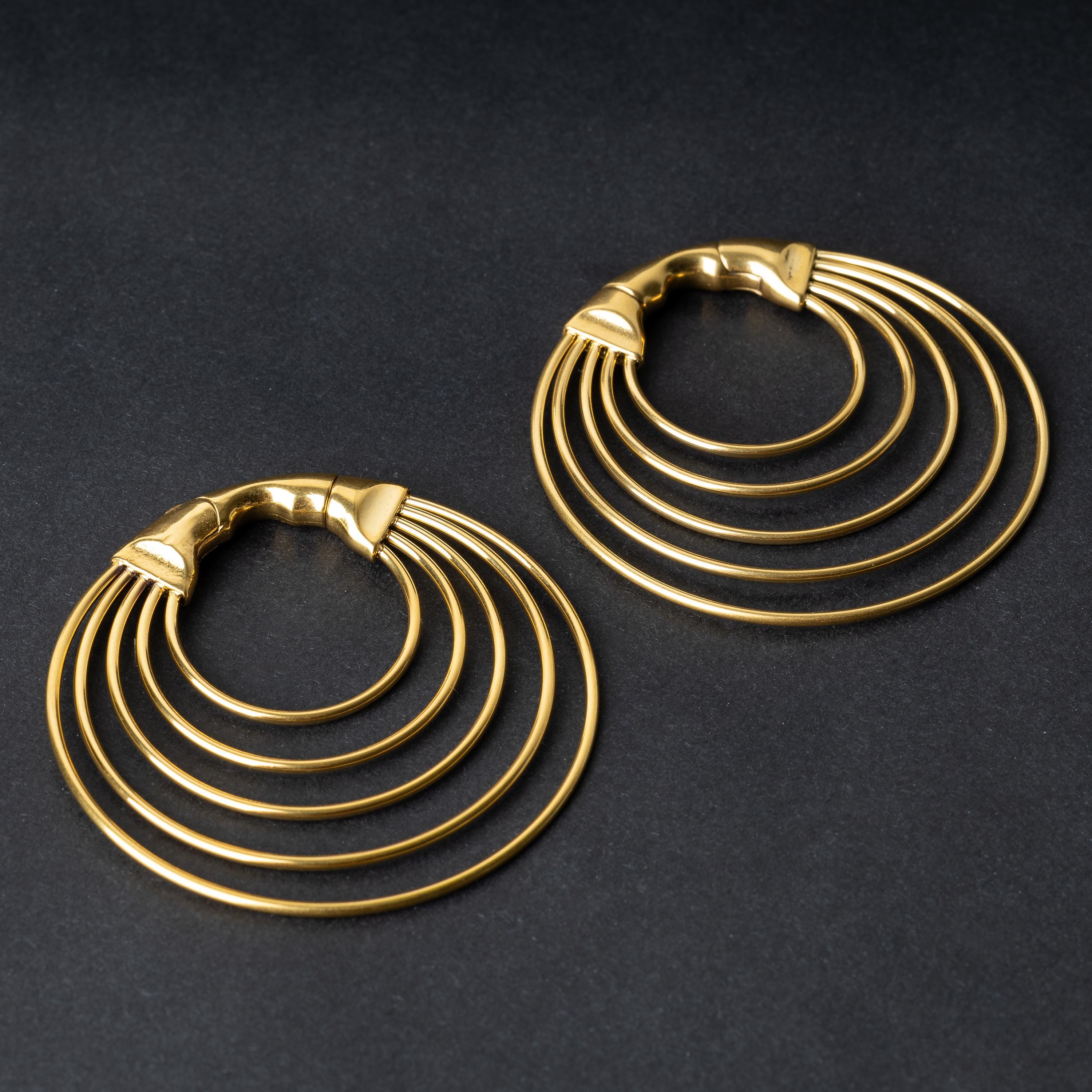 A pair of multi-hoop ear weights in polished gold, featuring five cascading hoops and a seamless hinge closure, displayed on a dark background.

