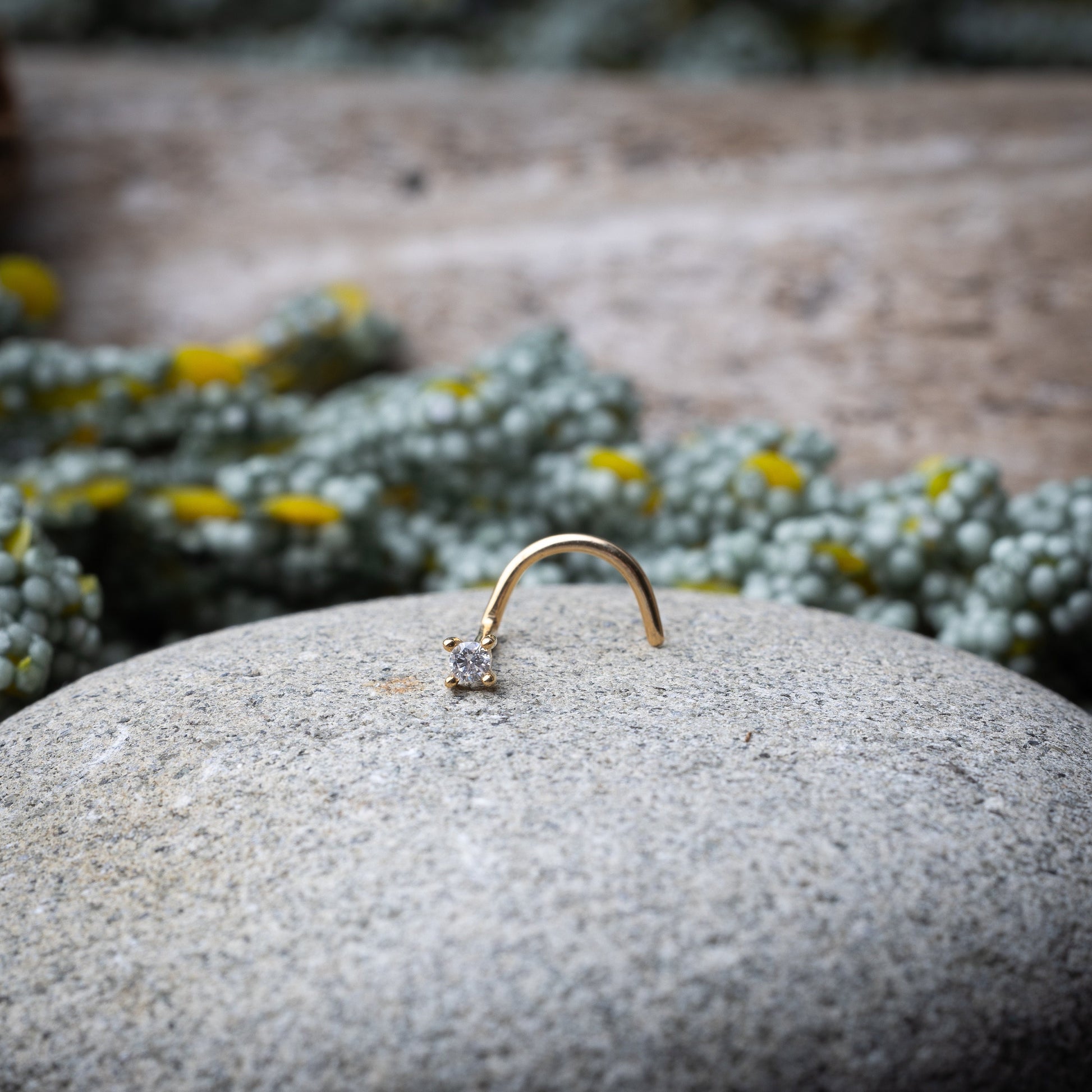 A gold-tone nose stud with a clear prong-set gem, featuring a curved design for secure wear, displayed on a stone background.