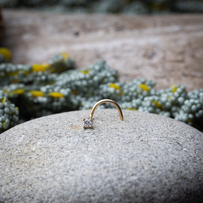 A gold-tone nose stud with a clear prong-set gem, featuring a curved design for secure wear, displayed on a stone background.