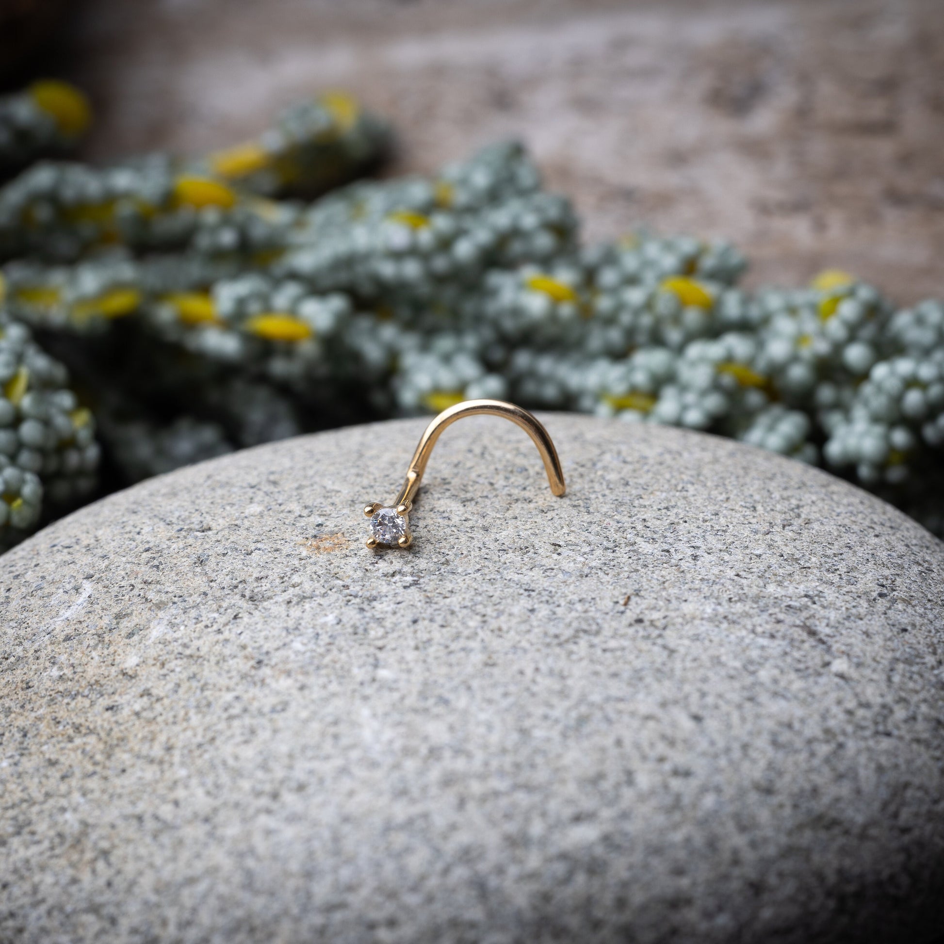 A gold-tone nose stud with a clear prong-set gem, featuring a curved design for secure wear, displayed on a stone background.