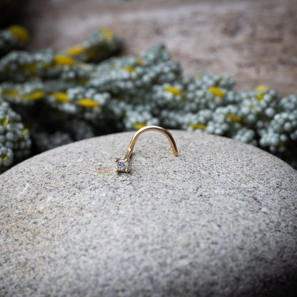 A gold-tone nose stud with a clear prong-set gem, featuring a curved design for secure wear, displayed on a stone background.