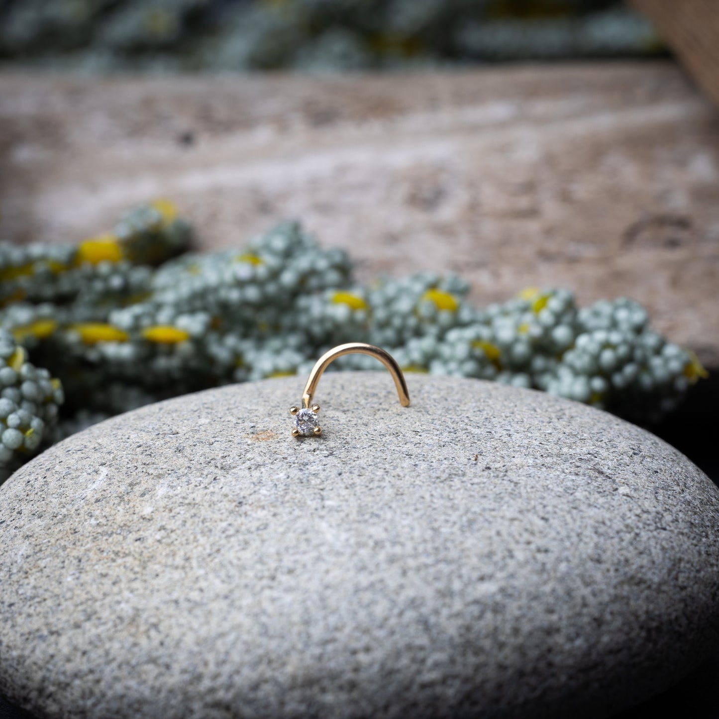 A gold-tone nose stud with a clear prong-set gem, featuring a curved design for secure wear, displayed on a stone background.
