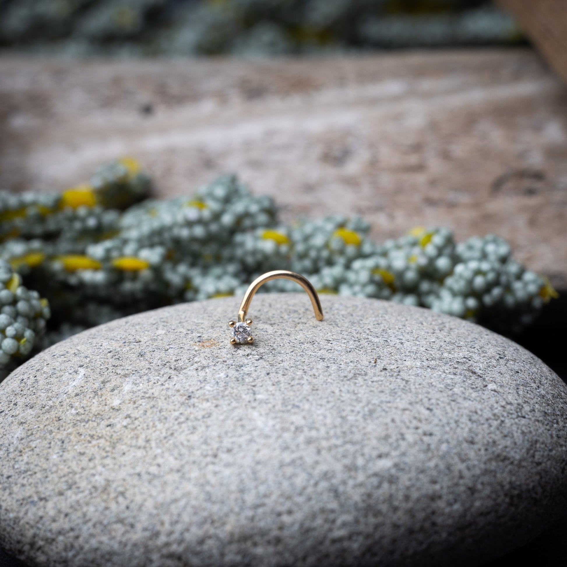 A gold-tone nose stud with a clear prong-set gem, featuring a curved design for secure wear, displayed on a stone background.