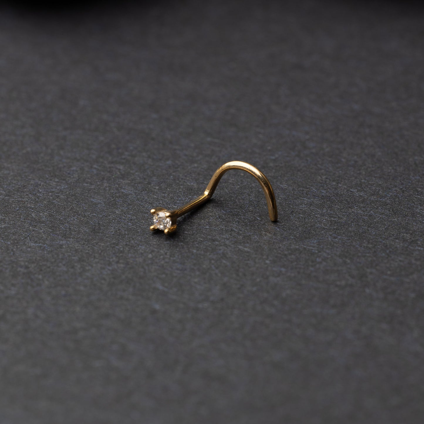 A gold-tone nose stud with a clear prong-set gem, featuring a curved design for secure wear, displayed on a dark background.