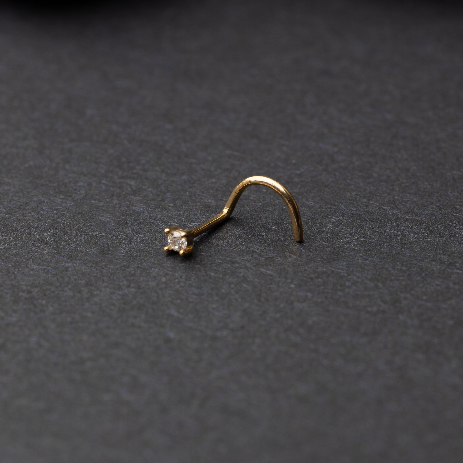 A gold-tone nose stud with a clear prong-set gem, featuring a curved design for secure wear, displayed on a dark background.