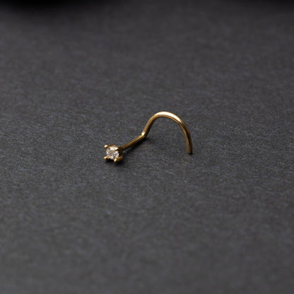 A gold-tone nose stud with a clear prong-set gem, featuring a curved design for secure wear, displayed on a dark background.