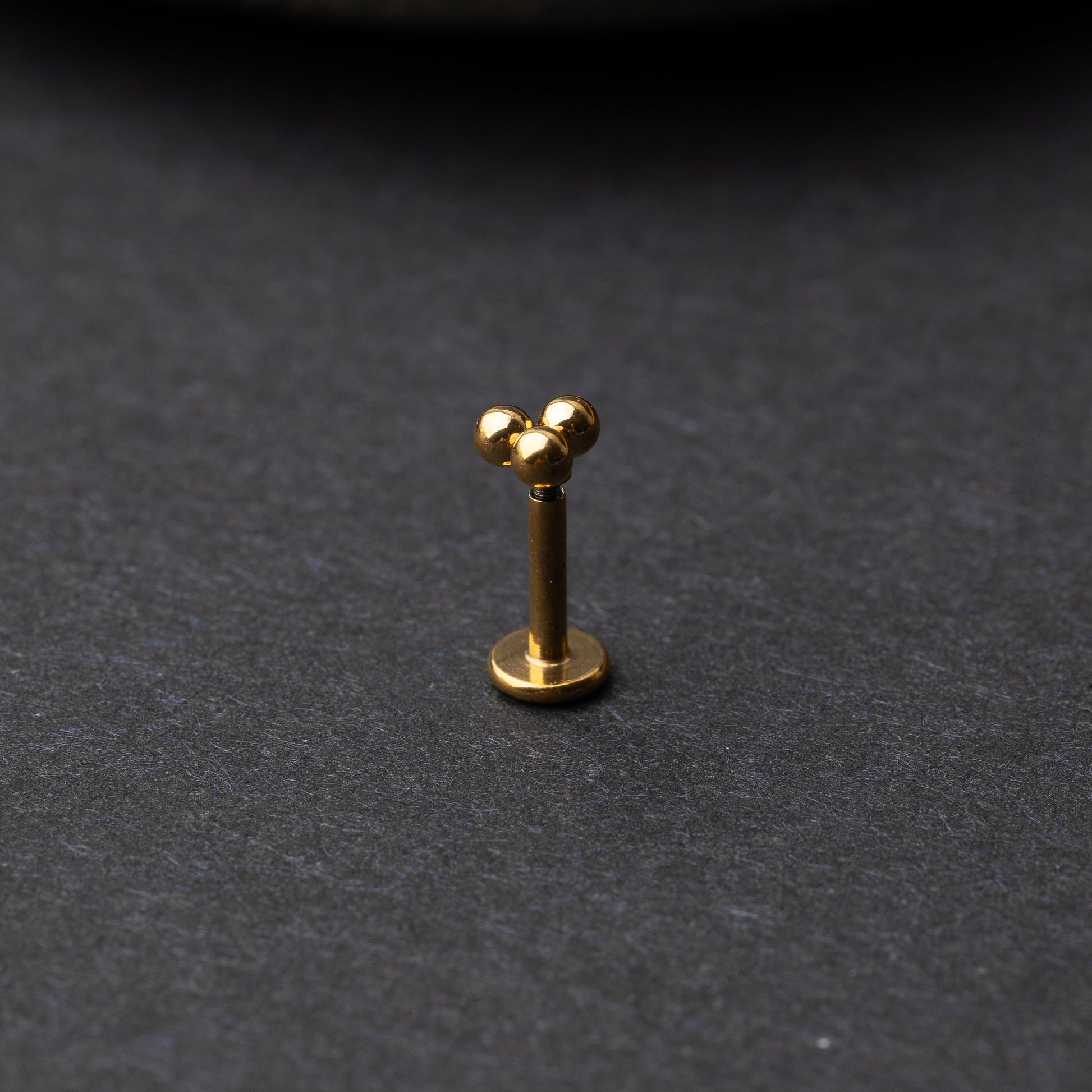 Close-up of three minimalist ball cluster labret studs in a gold finish, displayed on a dark background.

