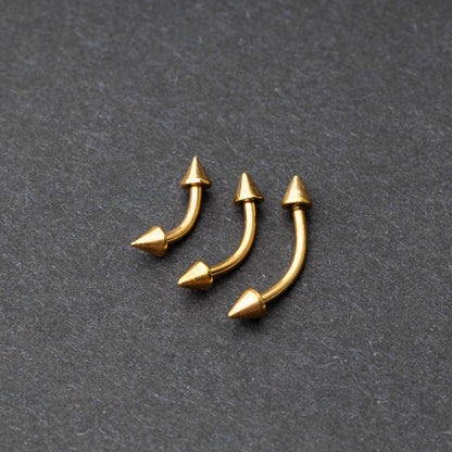Gold curved barbells with pointed spike ends, displayed on a dark background.