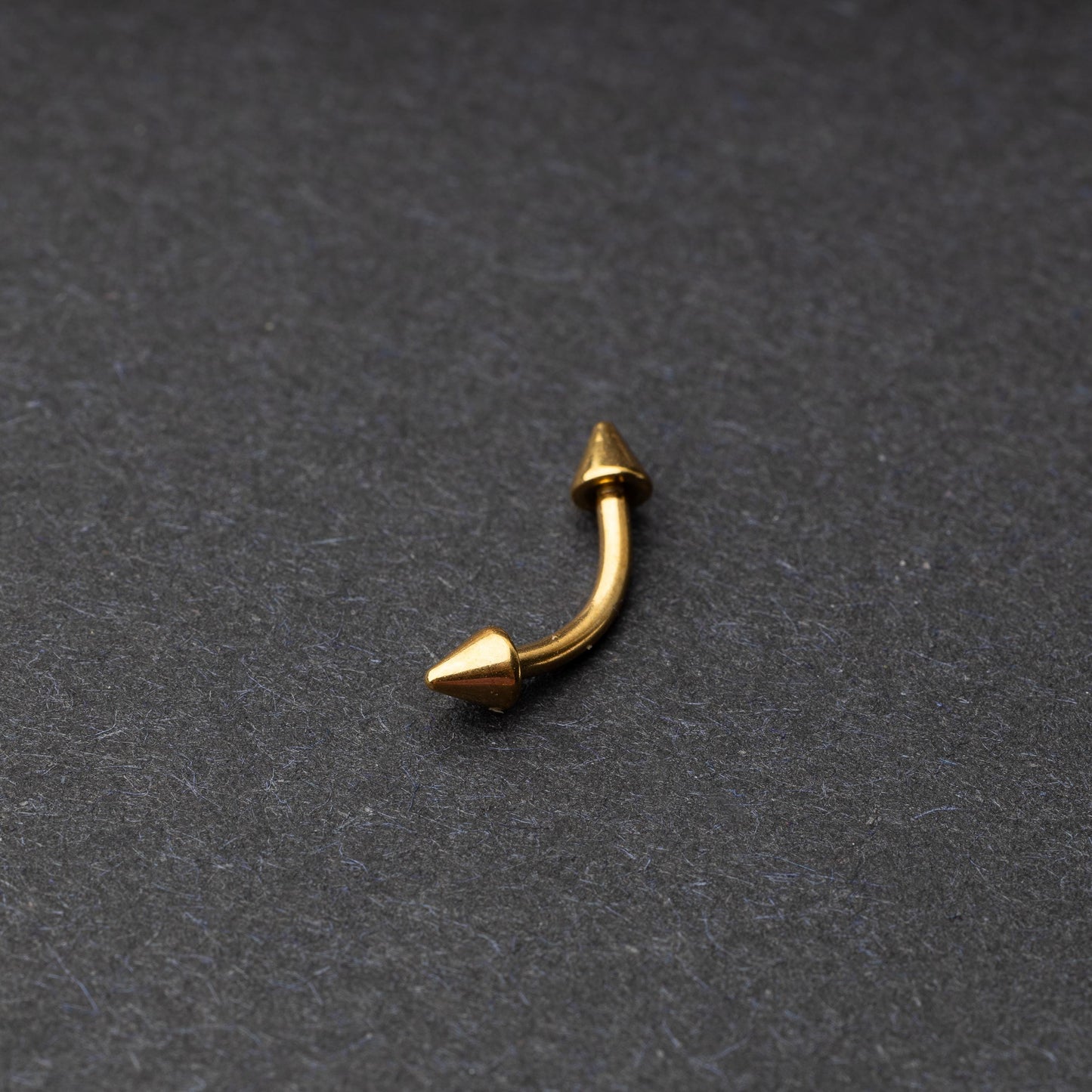 Gold curved barbell with pointed spike ends, displayed on a dark background.