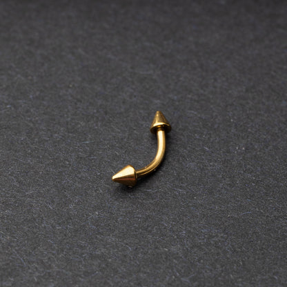 Gold curved barbell with pointed spike ends, displayed on a dark background.