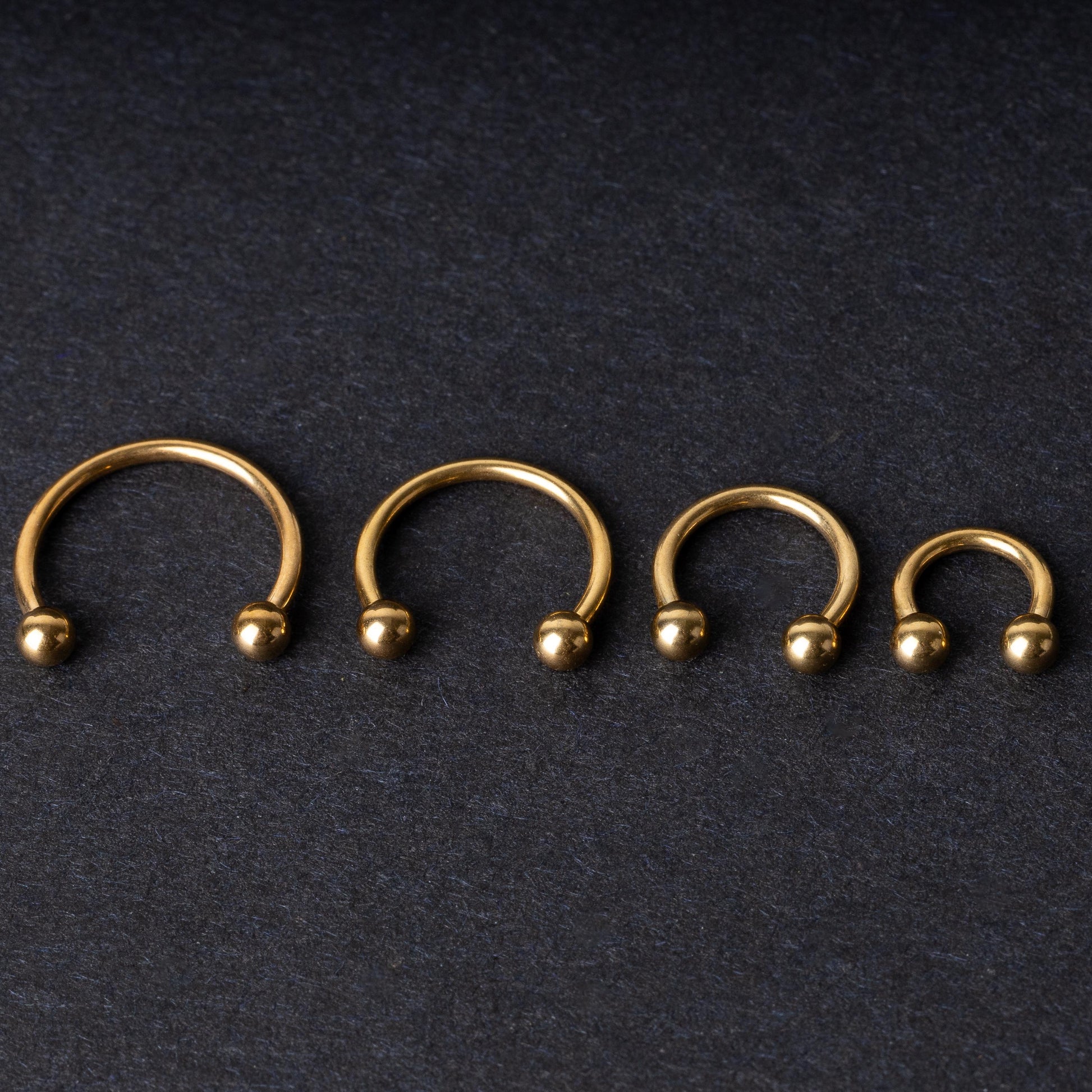Set of four gold-colored titanium horseshoe rings with ball ends, displayed on a dark textured background.

