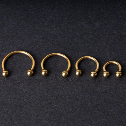 Set of four gold-colored titanium horseshoe rings with ball ends, displayed on a dark textured background.

