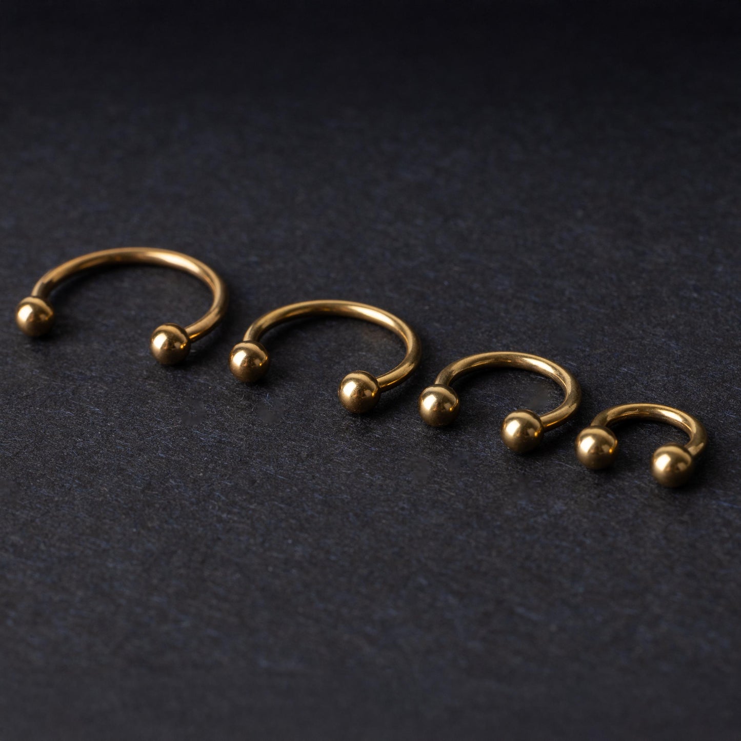 Set of four gold-colored titanium horseshoe rings with ball ends, displayed on a dark textured background.
