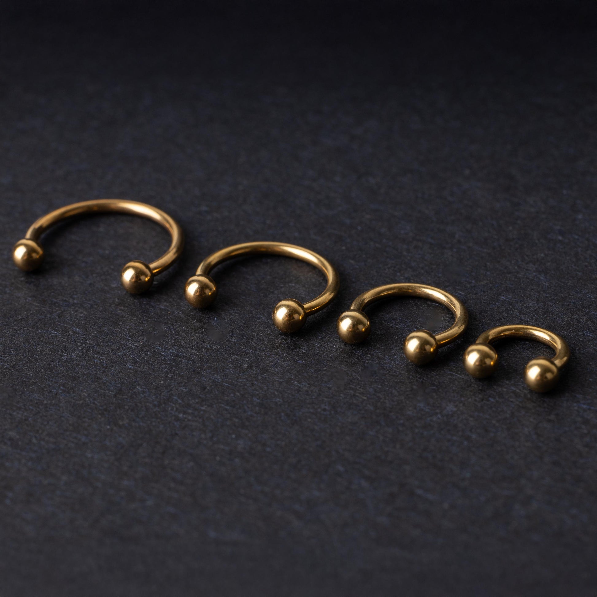 Set of four gold-colored titanium horseshoe rings with ball ends, displayed on a dark textured background.
