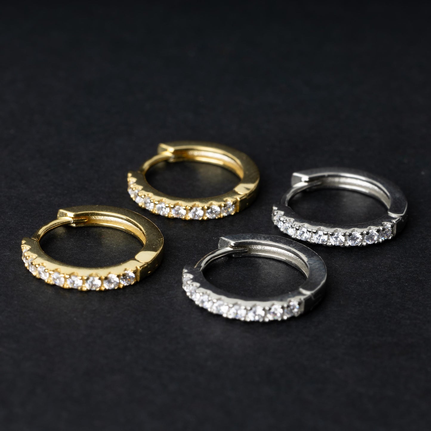 Two pairs of hoop earrings, one silver and one gold adorned with a row of sparkling cubic zirconia gems, displayed on a black background.