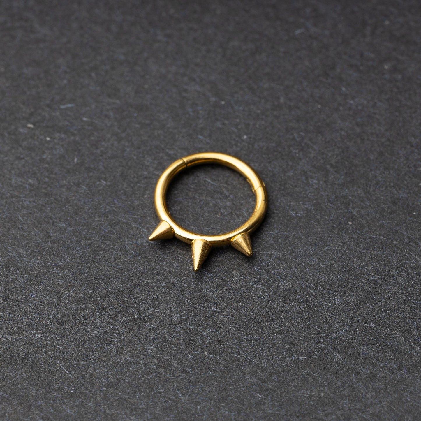 Gold-tone stainless steel clicker ring with three spikes, displayed on a dark textured background.
