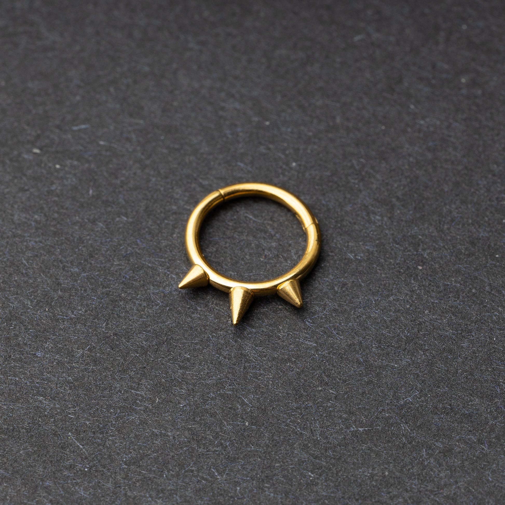 Gold-tone stainless steel clicker ring with three spikes, displayed on a dark textured background.