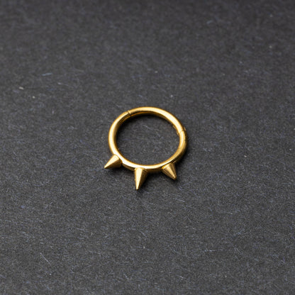 Gold-tone stainless steel clicker ring with three spikes, displayed on a dark textured background.