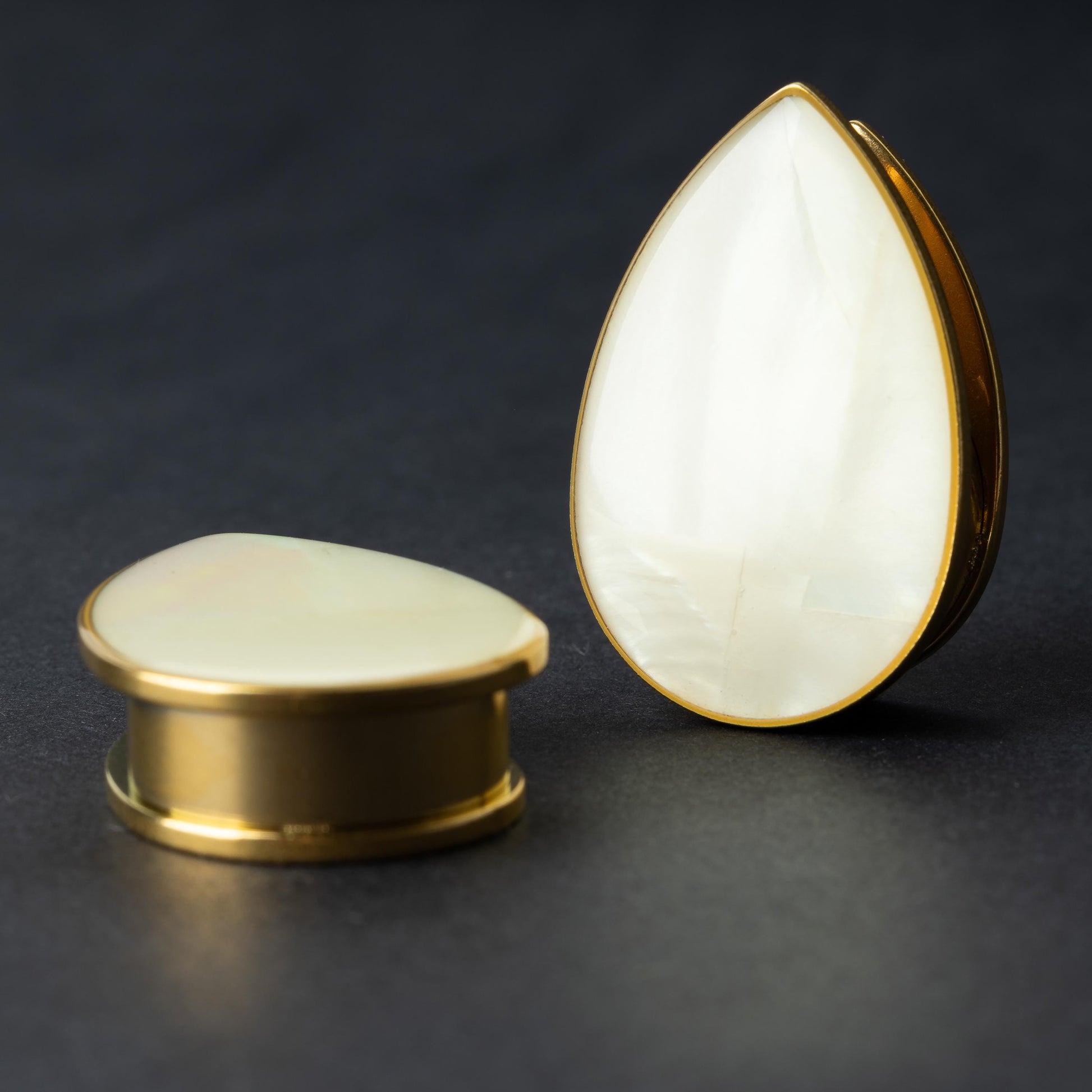 A pair of teardrop-shaped plugs with a polished mother-of-pearl inlay, framed by stainless steel, resting on a dark background.

