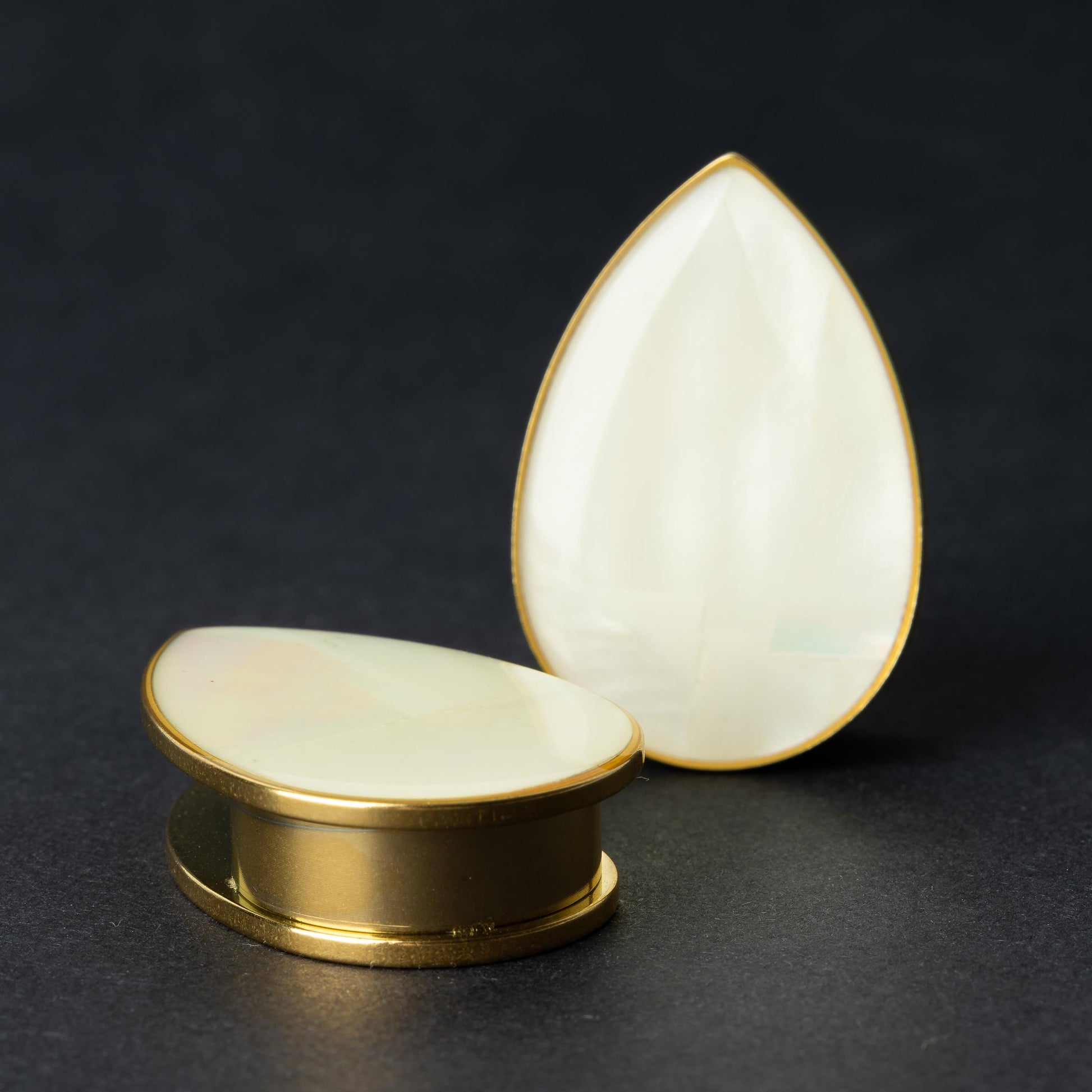 A pair of teardrop-shaped plugs with a polished mother-of-pearl inlay, framed by stainless steel, resting on a dark background.

