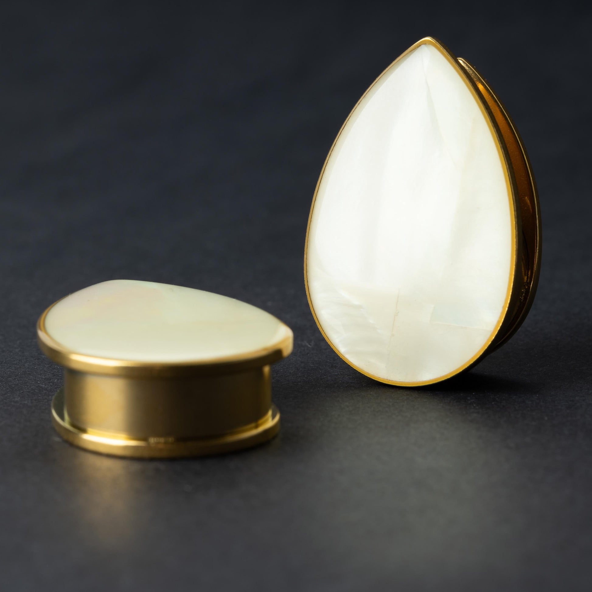 A pair of teardrop-shaped plugs with a polished mother-of-pearl inlay, framed by stainless steel, resting on a dark background.

