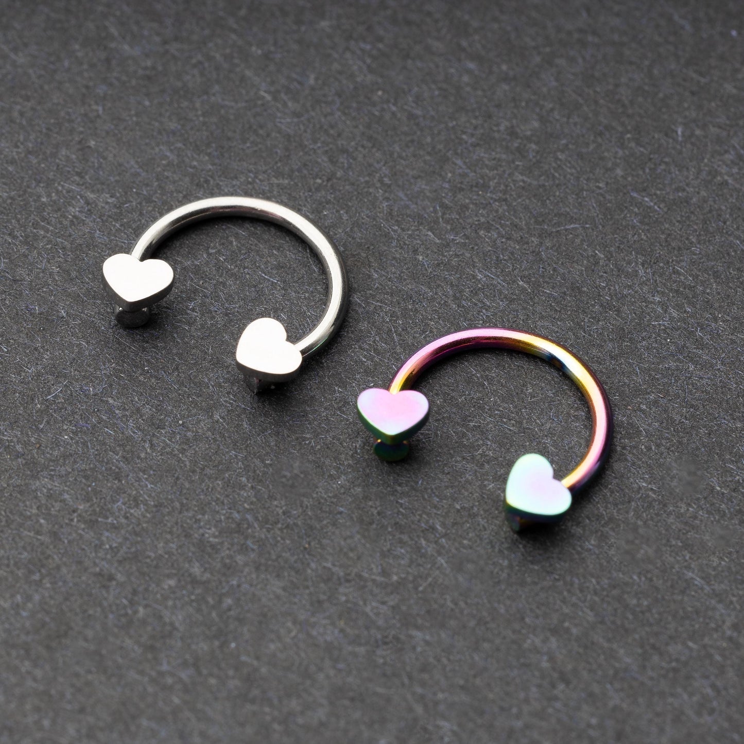 Two horseshoe-shaped hoops with heart-shaped ends in silver and rainbow finishes, displayed on a dark background.