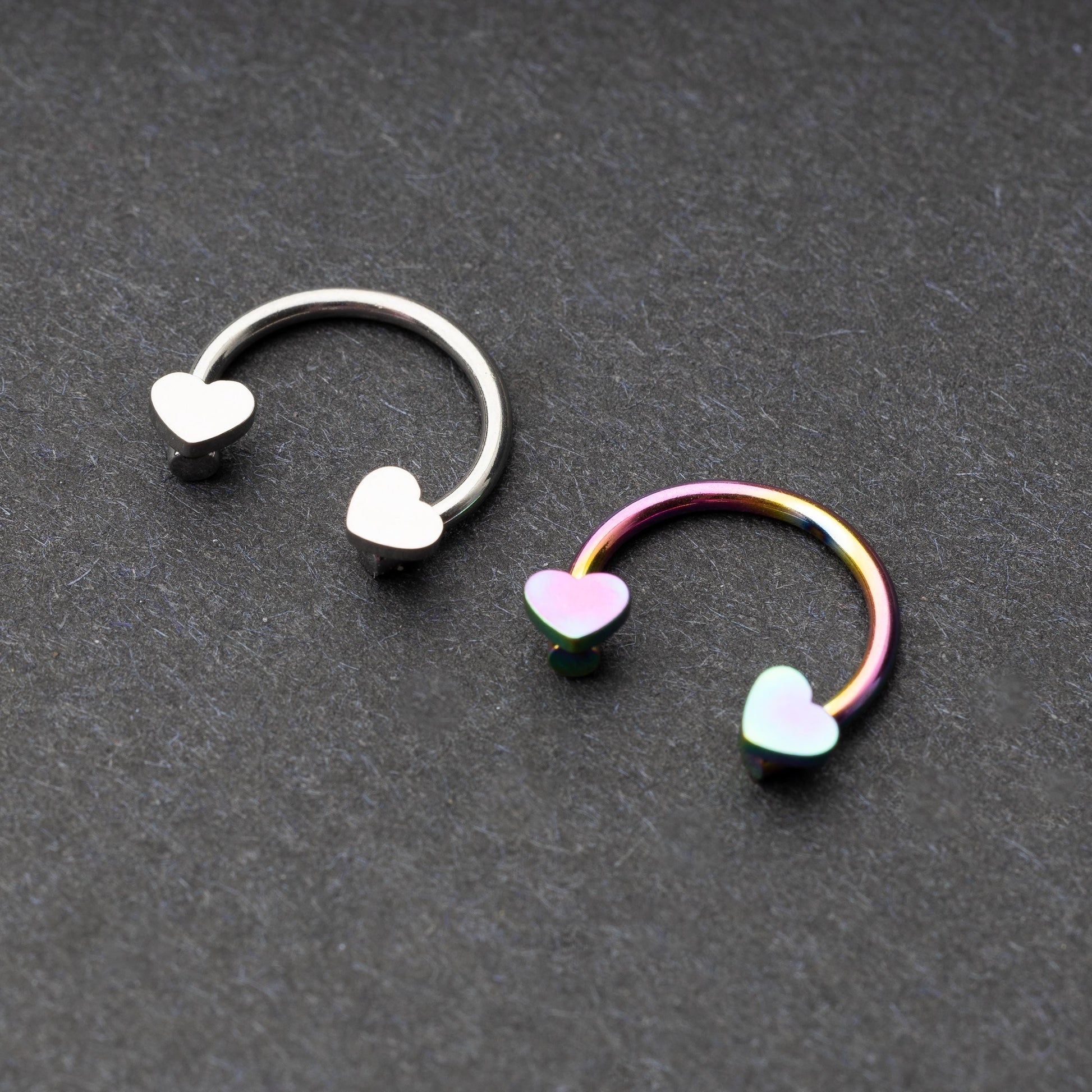 Two horseshoe-shaped hoops with heart-shaped ends in silver and rainbow finishes, displayed on a dark background.