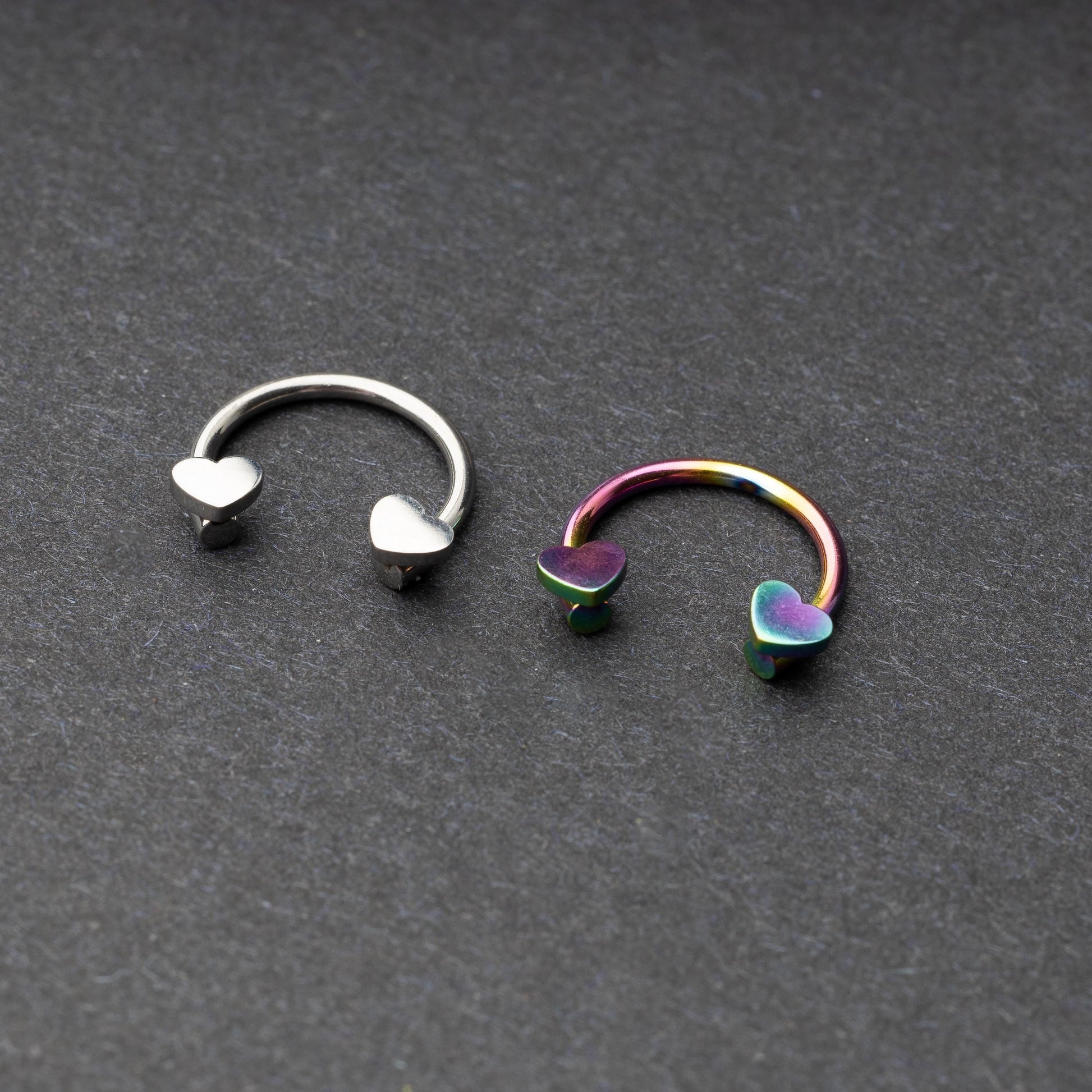 Two horseshoe-shaped hoops with heart-shaped ends in silver and rainbow finishes, displayed on a dark background.