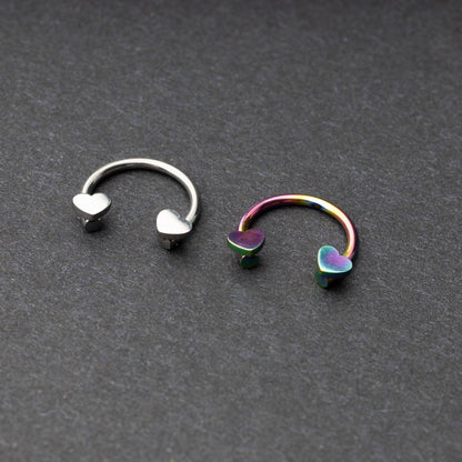 Two horseshoe-shaped hoops with heart-shaped ends in silver and rainbow finishes, displayed on a dark background.