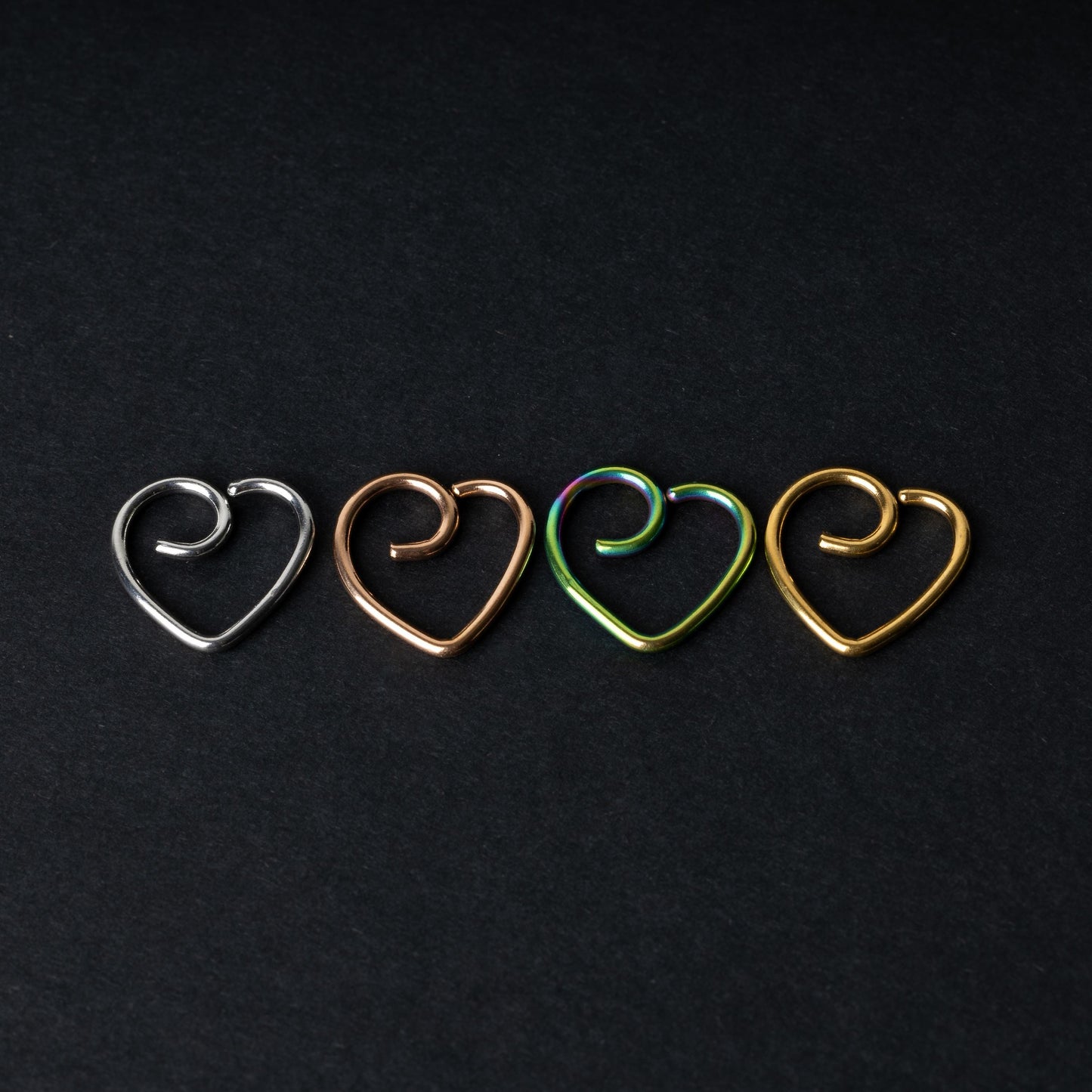 Four heart-shaped nose rings in silver, rose gold, rainbow, and gold finishes, displayed on a dark background.