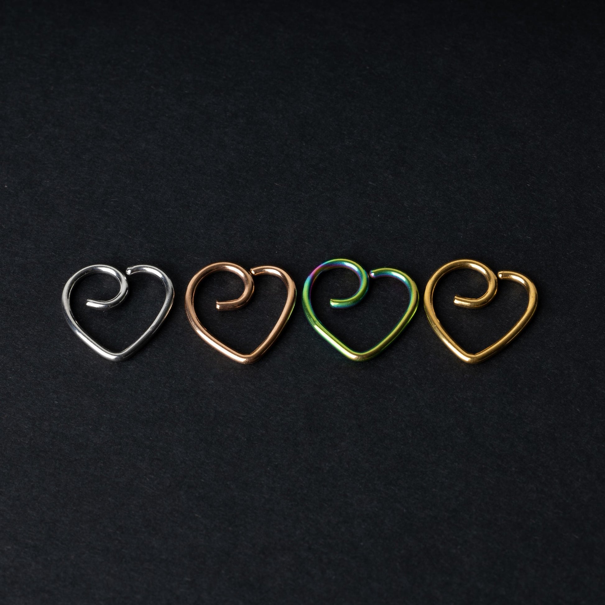 Four heart-shaped nose rings in silver, rose gold, rainbow, and gold finishes, displayed on a dark background.