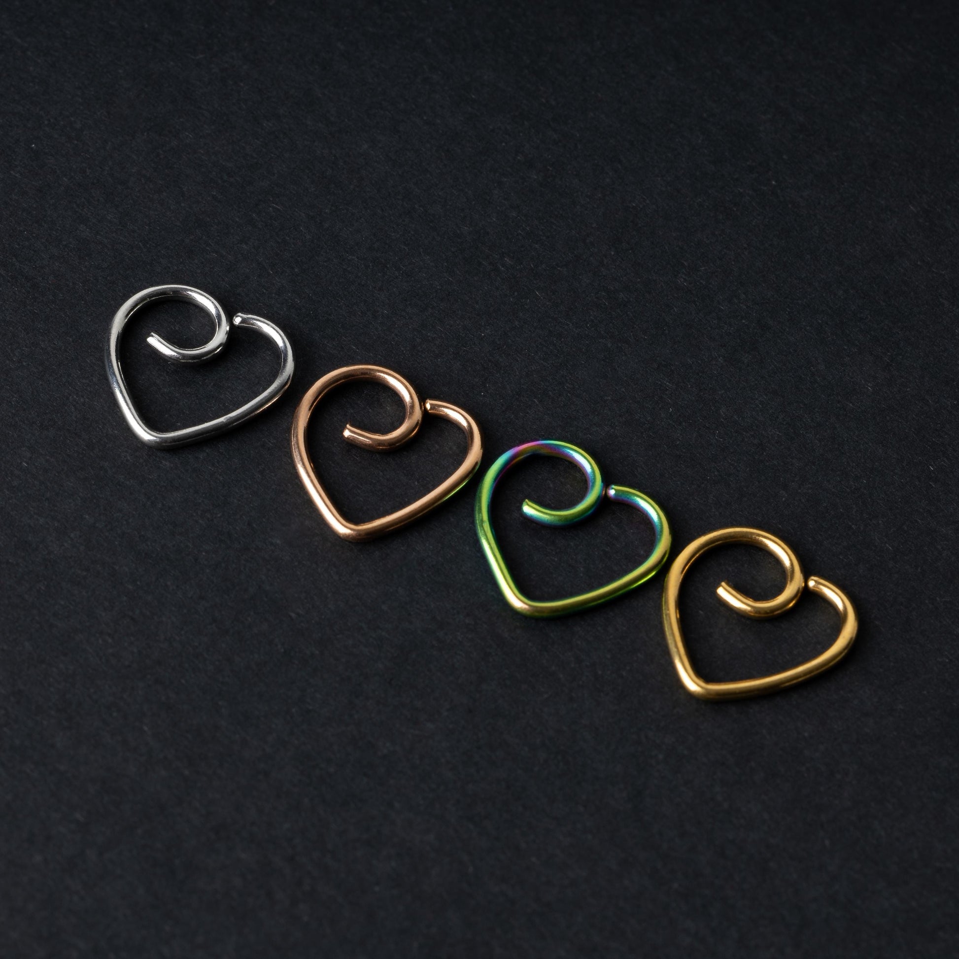Four heart-shaped nose rings in silver, rose gold, rainbow, and gold finishes, displayed on a dark background.
