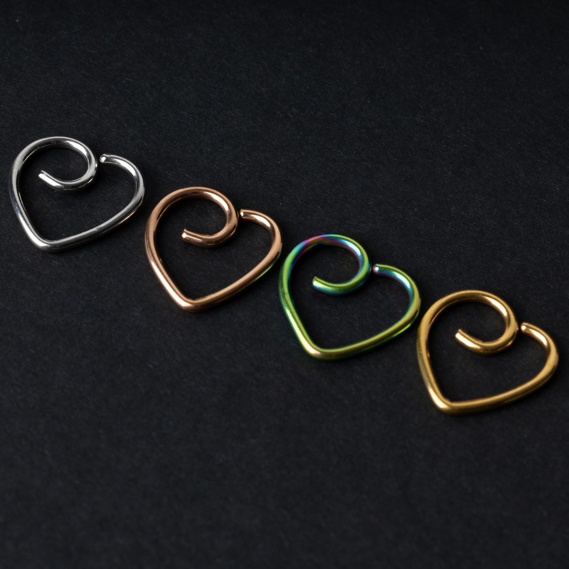 Four heart-shaped nose rings in silver, rose gold, rainbow, and gold finishes, displayed on a dark background.