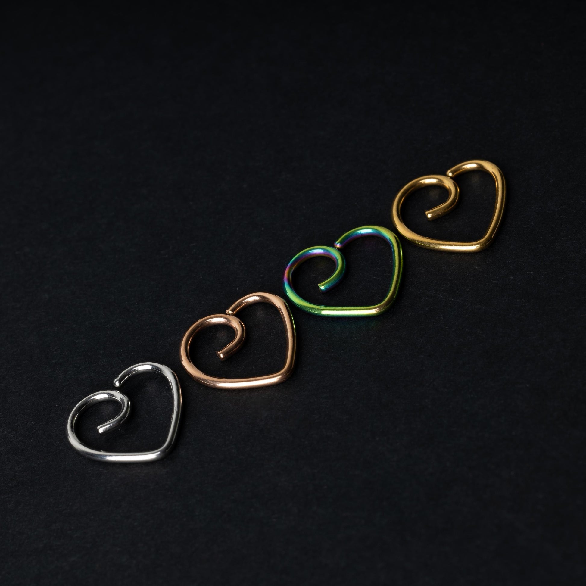 Four heart-shaped nose rings in silver, rose gold, rainbow, and gold finishes, displayed on a dark background.