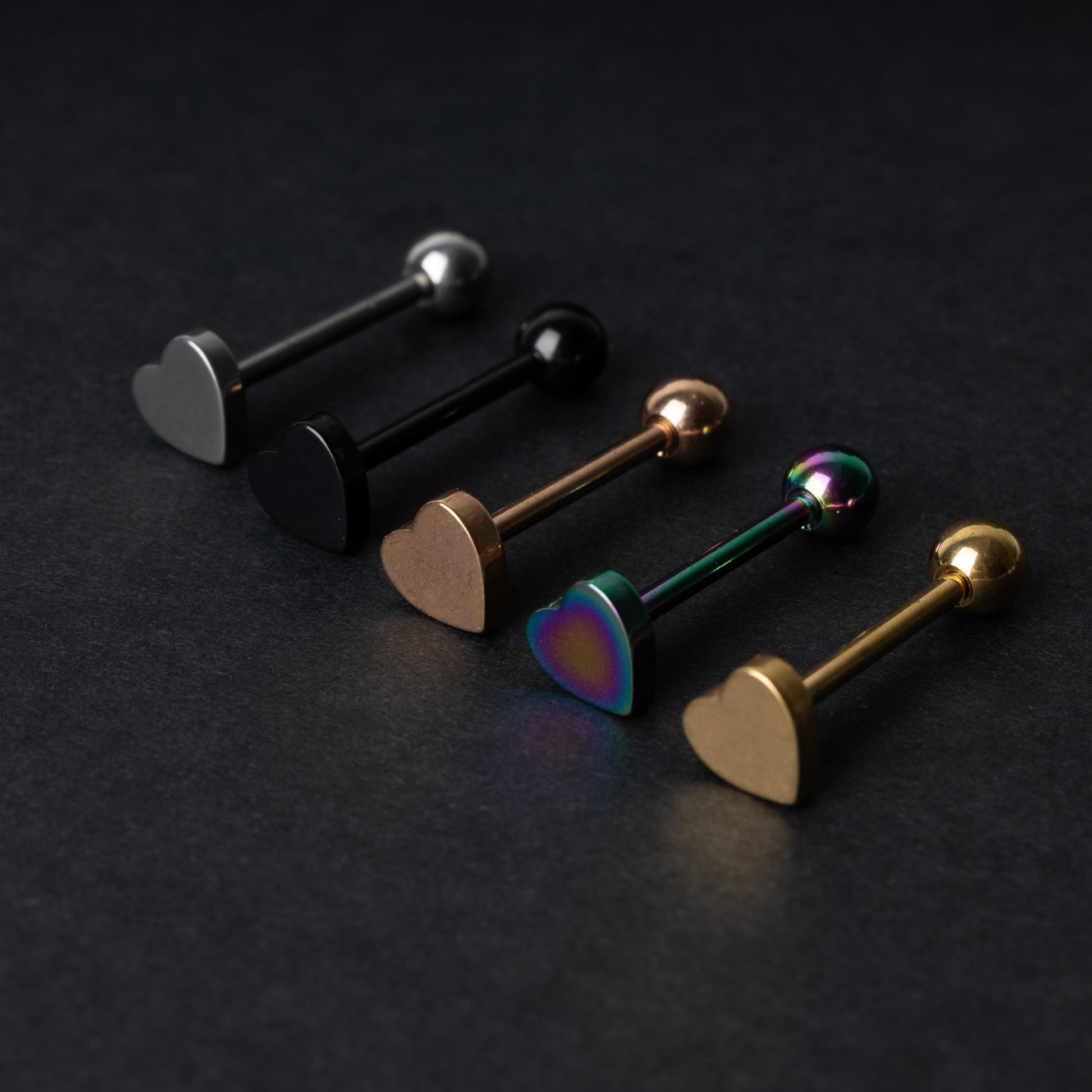 Heart-shaped tongue bars in black, silver, rose gold, rainbow, and gold finishes, displayed on a dark background.