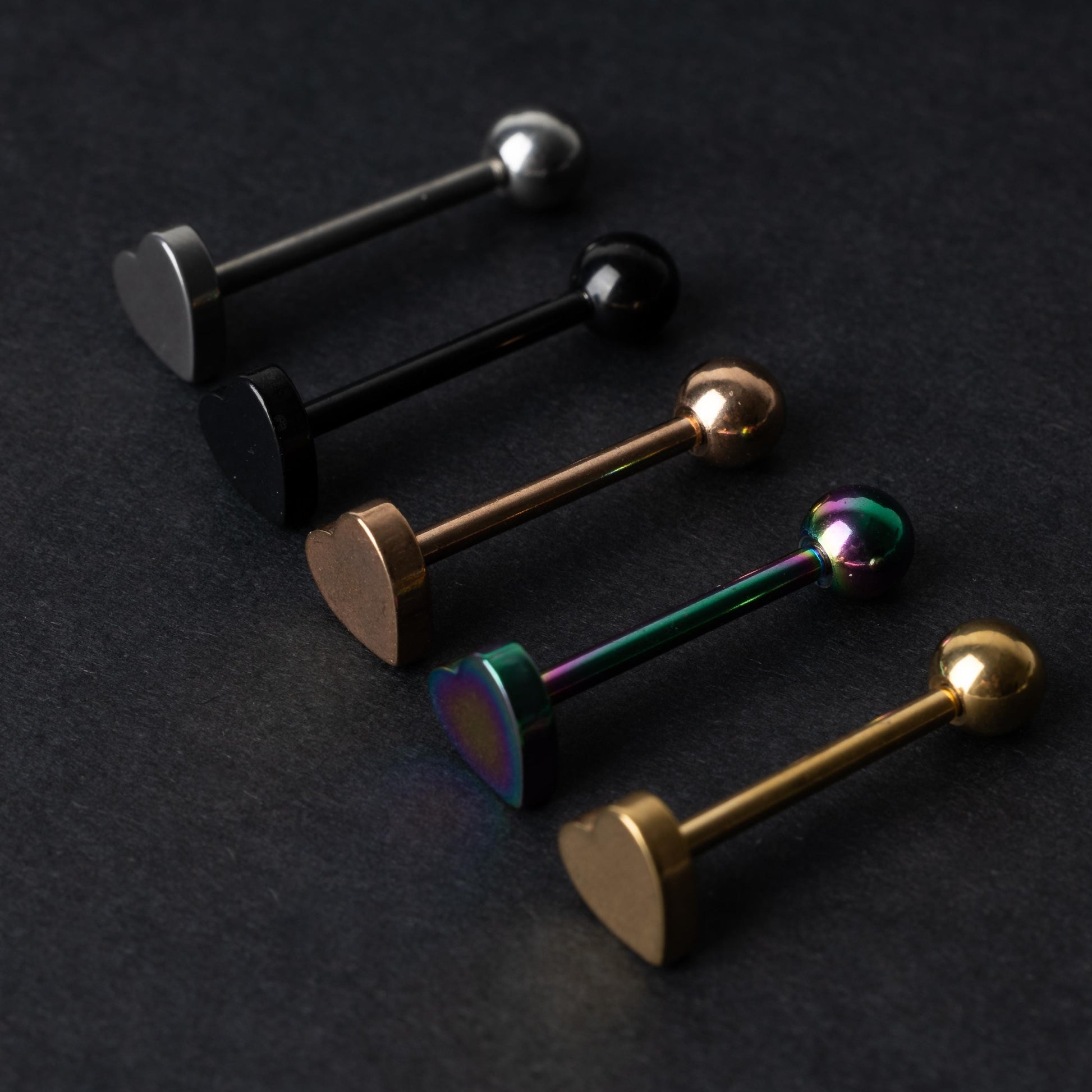 Heart-shaped tongue bars in black, silver, rose gold, rainbow, and gold finishes, displayed on a dark background.