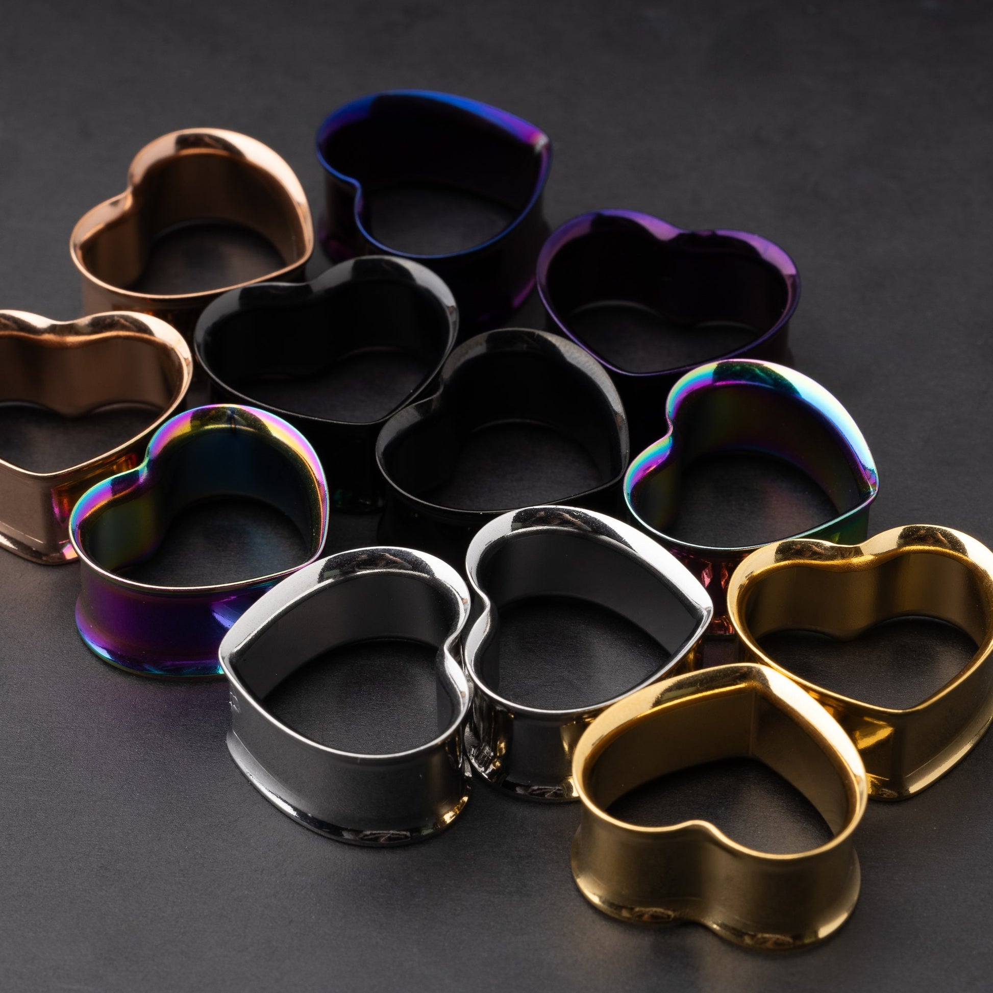 Heart-shaped tunnels in black, silver, rainbow, gold, purple, and rose gold finishes, displayed on a dark background.