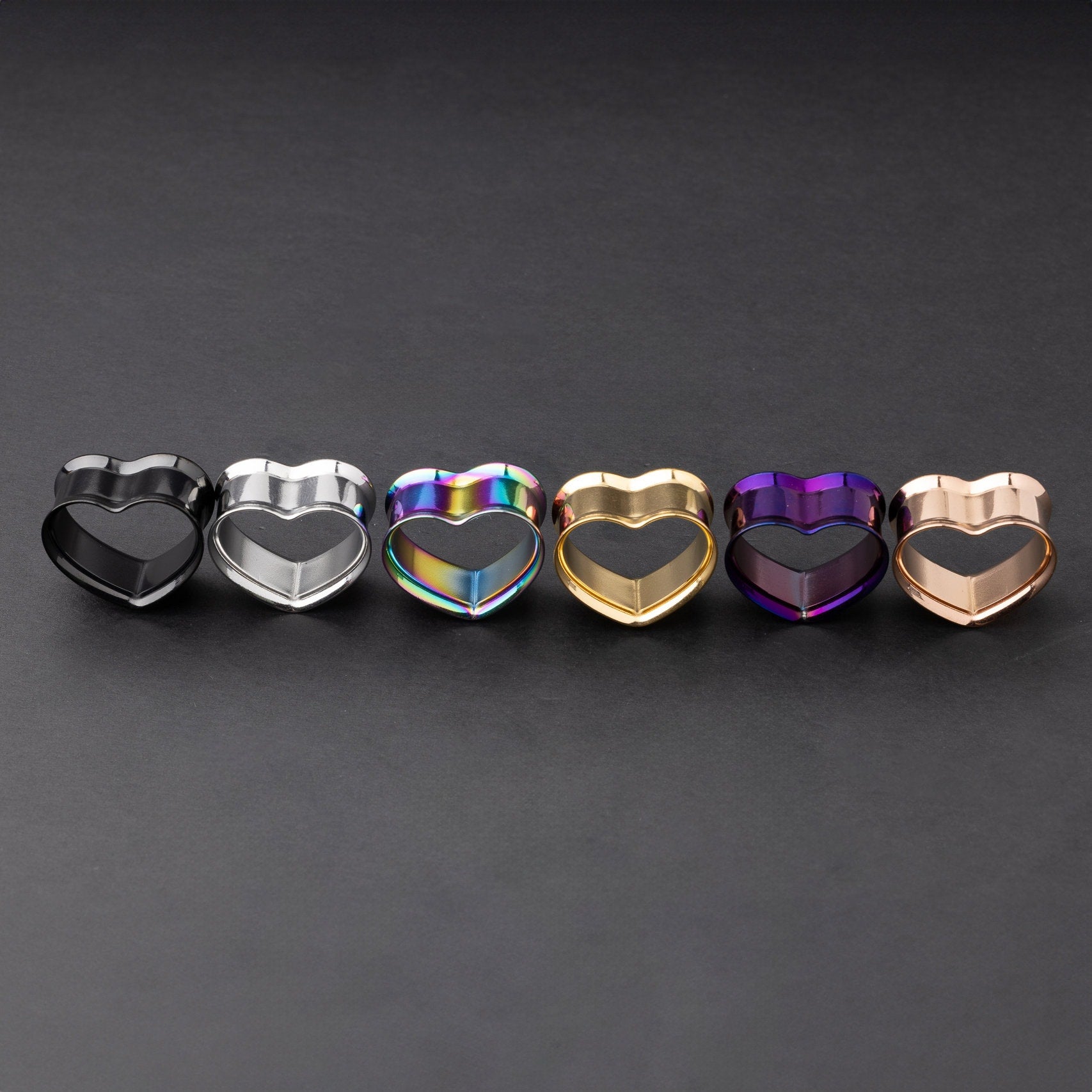 Heart-shaped tunnels in black, silver, rainbow, gold, purple, and rose gold finishes, displayed on a dark background.