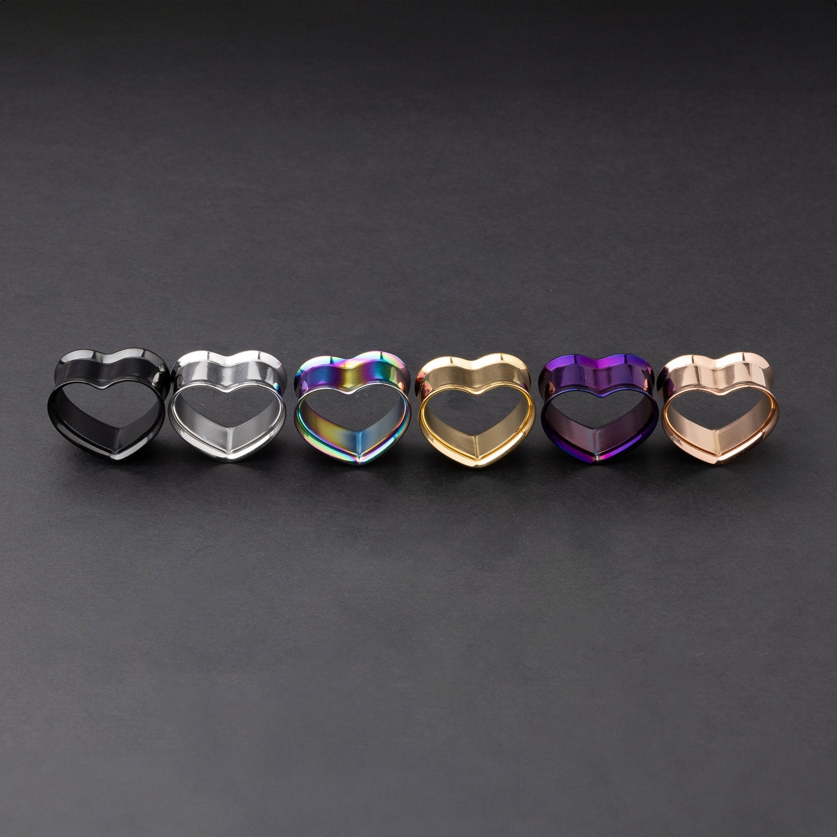 Heart-shaped tunnels in black, silver, rainbow, gold, purple, and rose gold finishes, displayed on a dark background.