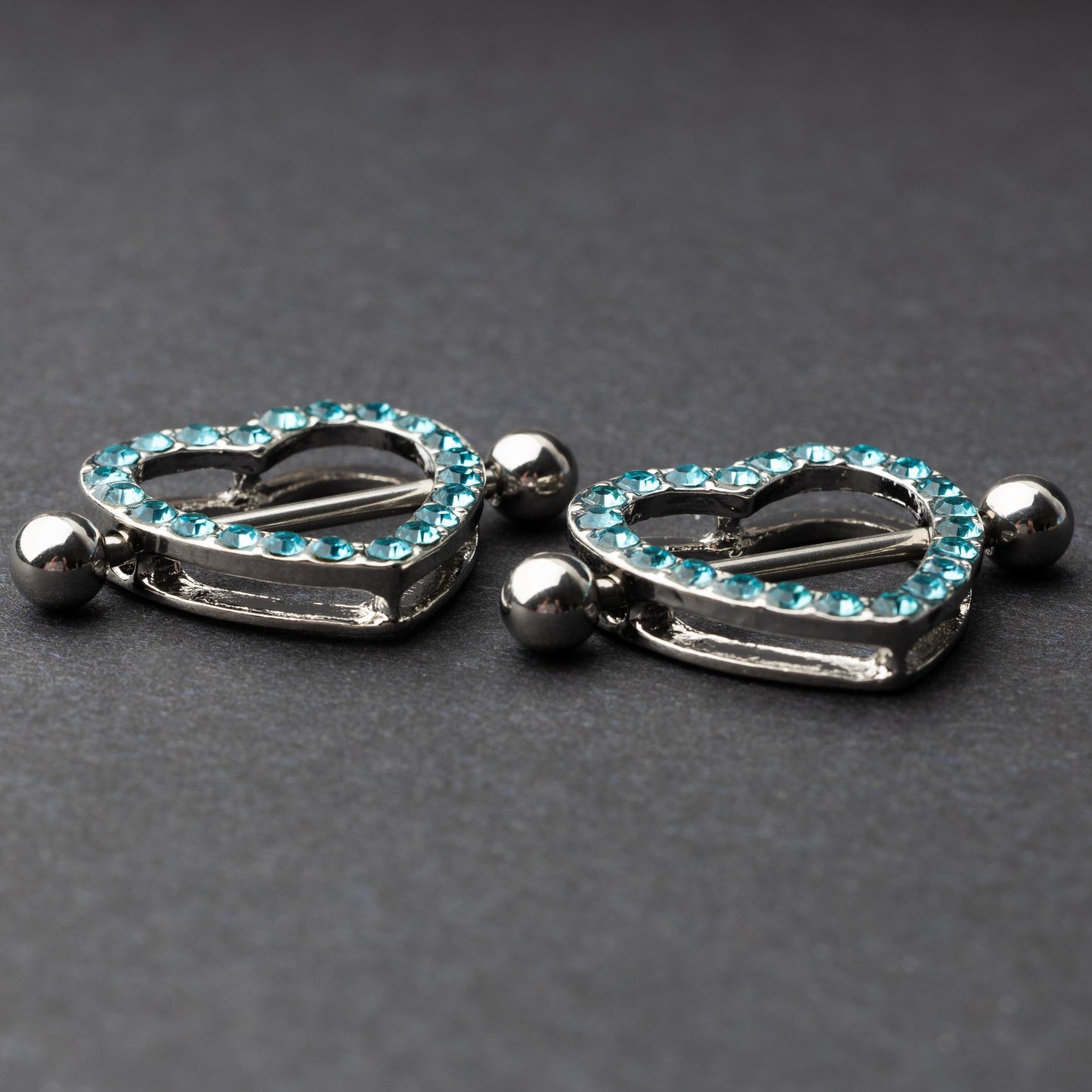 Two heart-shaped nipple barbells with silver metal and light blue gemstone accents, displayed on a dark background.