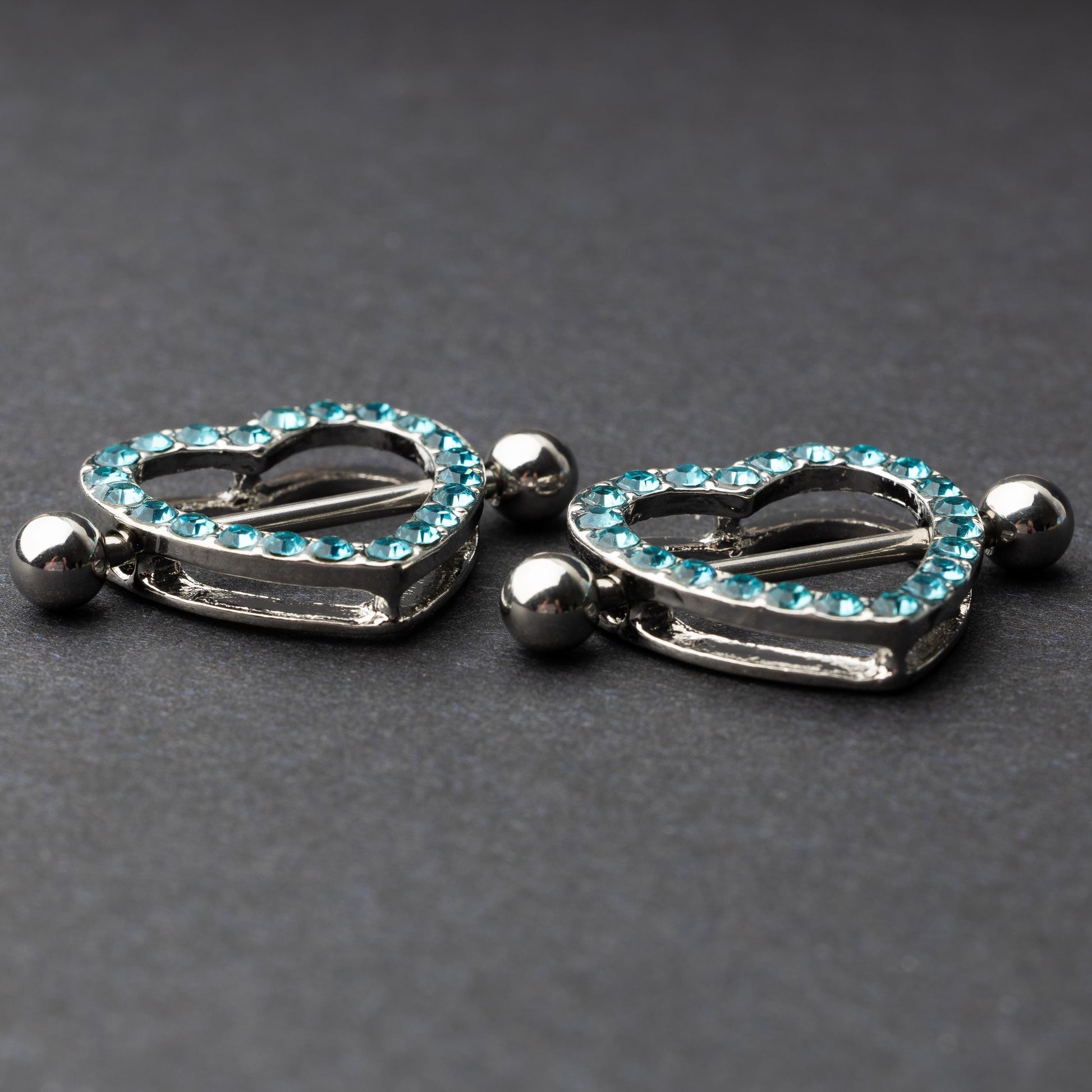 Two heart-shaped nipple barbells with silver metal and light blue gemstone accents, displayed on a dark background.